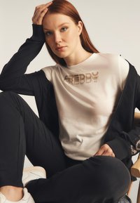 Camiseta blanca de manga corta con el logotipo metálico "FREDDY", combinada con una chaqueta negra con cremallera. Pantalones negros. Modelo sentado frente a un fondo claro.