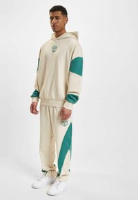 Beige hettegenser og matchende treningsthous med grønne detaljer; har logo trykk på brystet og buksene, med en avslappet passform og myk tekstur.