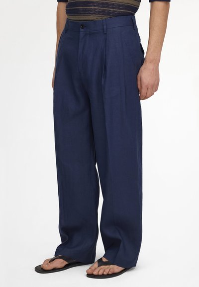 Homme portant un pantalon ample bleu marine et des tongs noires, debout les mains le long du corps devant un fond uni.