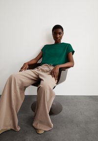Groene T-shirt met korte mouwen, gecombineerd met beige wijde broek. Model zit in een moderne stoel met de handen op de knieën. Zachte texturen en een eenvoudig ontwerp.