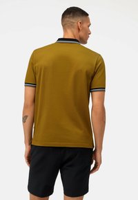 Kurzarm-Poloshirt in Olivgrün mit schwarzem Kragen und doppelten Streifenakzenten. Getragen mit schwarzen Shorts, die ein Schmetterlingstattoo am Arm zeigen.