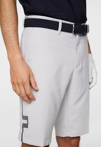 Ljusgrå golfshorts gjorda av slät tyg, med ett svart flätat bälte och en randig detalj vid fåll.