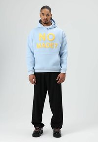 Sweat à capuche bleu clair avec texte brodé jaune "NO MADE?", doté d'une poche avant. Associé à un pantalon noir ample et des chaussures noires.