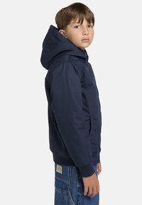 Námořnický modrý mikina s kapucí vyrobená z odolné látky, s předním zipem, bočními kapsami a žebrovanými manžetami. Model má na sobě volné džíny.
