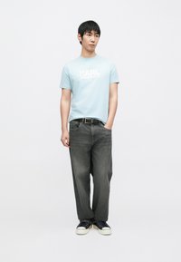 Világoskék póló fehér "KARL LAGERFELD" felirattal, laza, sötétszürke farmernadrággal és fekete sneaker cipőkkel, fehér talppal. Egyszerű dizájn.