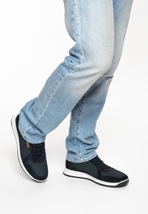 Light blue jeans and black perforated sneakers with white soles worn on crossed legs against white background.