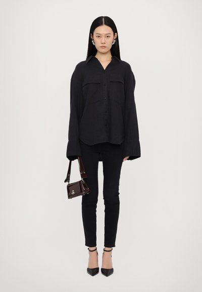 Black long-sleeve shirt with button-down front, two chest pockets, paired with black skinny jeans and pointed-toe black heels. Holding a small dark handbag.