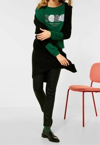 Grön sweatshirt med en glittrande grafik och text, lager på lager med en svart strukturerad sjal. Svarta vadderade byxor, gröna strumpor och svarta loafers.