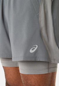 Grå træningsshorts med en glat overflade og ribbet inderside. Har et logo på nederste venstre side. Inkluderer sidelommer og syningsdetaljer.