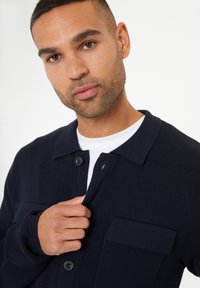 Námořnická modrá pletená kardigan s límcem, zapínáním na knoflíky a náprsní kapsou. Textura látky je žebrovaná a střih je volný.