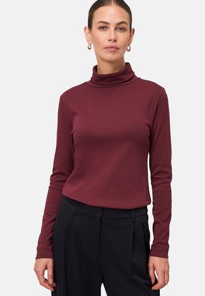 Femme portant un pull à col roulé bordeaux à manches longues rentré dans un pantalon noir taille haute avec des boucles d'oreilles créoles, se tenant devant un fond uni.
