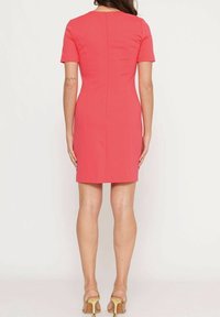 Žena v těsném korálově růžovém krátkém rukávu šatech a béžových vysokých podpatkách, stojící zády k fotoaparátu na jednobarevném pozadí.