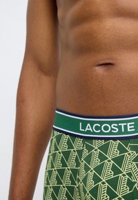 Πράσινα ανδρικά εσώρουχα μποξέρ με γεωμετρικό σχέδιο σε αποχρώσεις του πράσινου και κίτρινου. Η ελαστική ζώνη έχει τη λέξη "LACOSTE" σε λευκό και σκούρο μπλε.