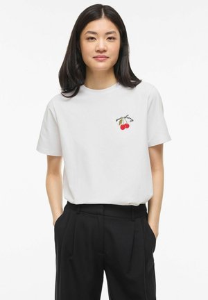 Ung kvinde med langt mørkt hår iført en hvid t-shirt med en lille kirsebærgrafik og sort højtaljede bukser, hænderne i lommerne.