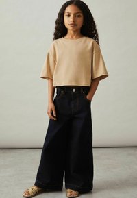 Beige cropped t-shirt med korte ærmer, parret med mørkeblå vidde jeans med kontrasterende orange syninger og frontlommer. Bæres med brune sandaler.