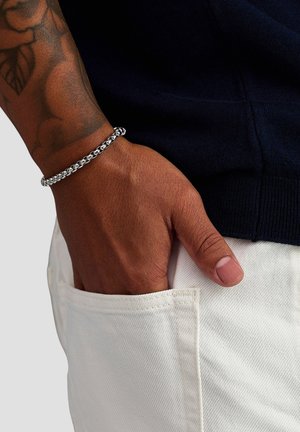 Silver chain bracelet with interlocking links, worn on a hand resting in a white denim pocket. Dark fabric in the background.