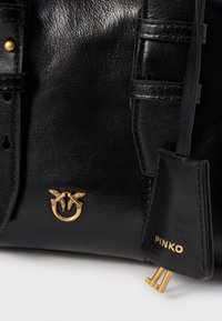 Μαύρη δερμάτινη τσάντα με χρυσό έμβλημα πουλιού και χρυσή ετικέτα "PINKO" προσαρτημένη σε μία θήκη.