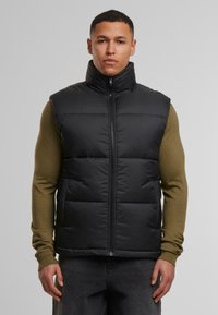 Schwarze gepolsterte Weste mit hohem Kragen, Reißverschluss vorne und gestepptem Design. Getragen über einem olivgrünen Langarmoberteil und grauen Hosen.