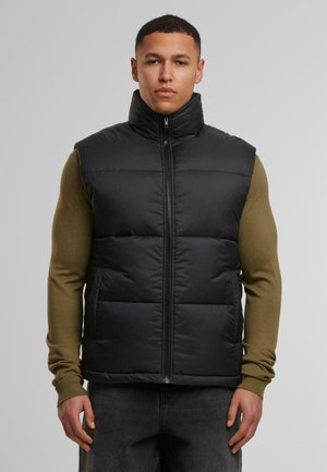 Schwarze gepolsterte Weste mit hohem Kragen, Reißverschluss vorne und gestepptem Design. Getragen über einem olivgrünen Langarmoberteil und grauen Hosen.