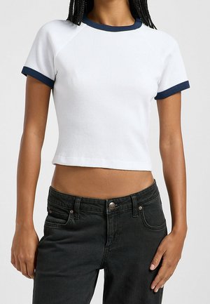 Persona que lleva una camisa blanca corta de manga corta con ribete azul marino y jeans negros de talle alto con un botón visible.