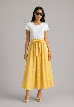 Femme portant des lunettes de soleil noires, une chemise blanche à manches courtes, une jupe jaune à pois avec un nœud, et des talons ouverts beige, debout devant un fond gris.