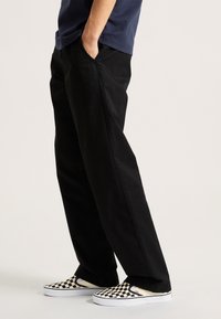 Black cotton trousers with a straight-leg design, worn with checkerboard slip-on shoes featuring a white sole and contrasting black accents.
