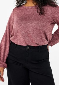 Una donna che indossa un top lungo a maniche con texture color bordeaux, infilato in pantaloni neri a vita alta, con una mano sull'anca e l'altra rilassata.