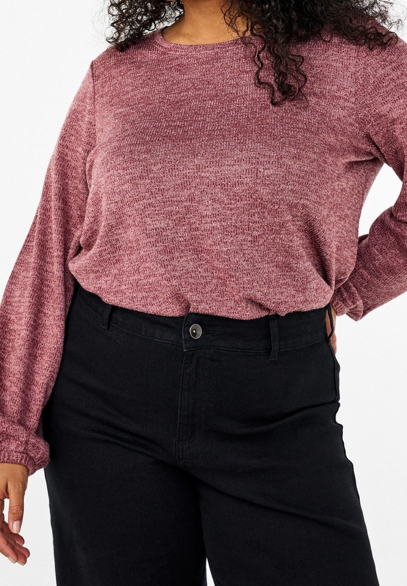 Una donna che indossa un top lungo a maniche con texture color bordeaux, infilato in pantaloni neri a vita alta, con una mano sull'anca e l'altra rilassata.