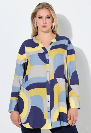 Kobieta w długim, zapinanym na guziki tuniku z abstrakcyjnymi, zakrzywionymi wzorami w kolorach niebieskim, żółtym, szarym i granatowym, stojąca na białym tle.