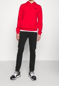 Czerwona bluza z kapturem z przednią kieszenią oraz logo, zestawiona z czarnymi spodniami o wąskim kroju i czarno-białymi sneakersami.