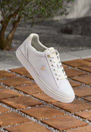 Zapatilla de cuero blanca con una textura suave, punta redonda, diseño con cordones y acento del logo dorado. Colocada sobre baldosas de madera y grava.