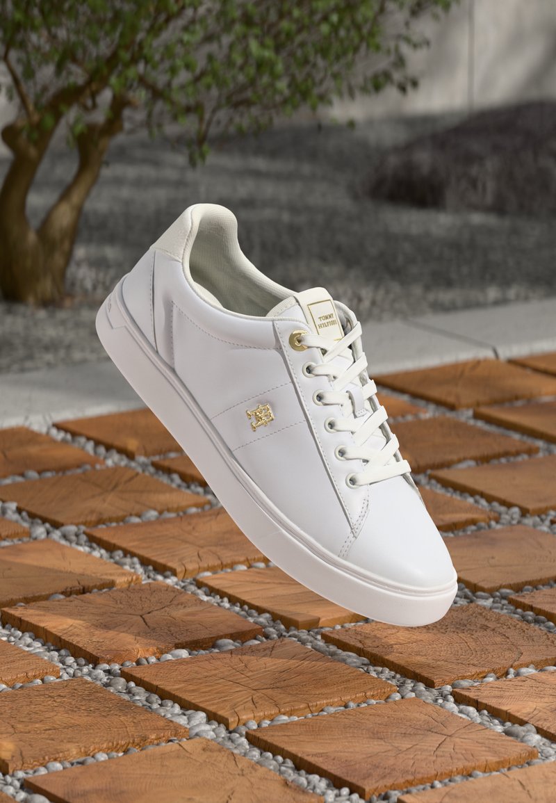 S sapatilhas brancas de couro, com uma textura suave, bico arredondado, design de atacadores e um detalhe em logotipo dourado. Colocadas sobre azulejos de madeira e cascalho.