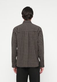 Un uomo che indossa una camicia a quadri marrone e nera con maniche lunghe, girato di spalle, abbinata a pantaloni neri su uno sfondo bianco uniforme.