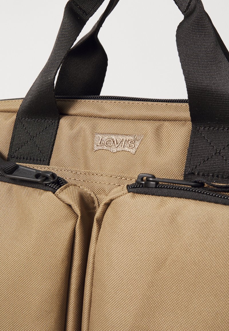 Beige Stofftasche mit doppeltem Reißverschluss, schwarzen Akzenten und einem auffälligen gestickten Levi's-Logoetikett. Robuste Griffe für bequemen Transport.