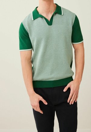 Homme portant un polo à manches courtes à rayures vertes et blanches avec un pantalon sombre, debout devant un fond uni clair.