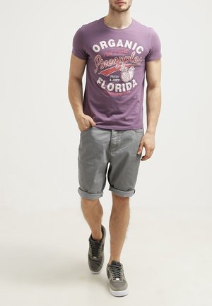 Camiseta morada con gráfico "Organic Pineapples Florida", combinada con pantalones cortos grises con dobladillo enrollado y zapatillas deportivas grises.