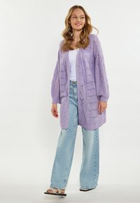 Lila stickad cardigan med öppen framtill, långa ärmar och intrikata mönsterdetaljer. I kombination med ljusblå vida jeans och sandaler.
