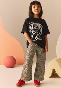 Czarna koszulka z grafiką Rolling Stones, szare spodnie w panterkę o szerokich nogawkach i czerwone sneakersy. Tło przedstawia miękkie, zakrzywione pastelowe kształty.