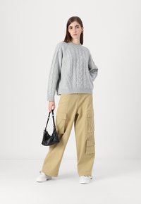Sivkasta pletena jopica z širokimi rebrastimi detajli, v kombinaciji z khaki cargo hlačami, ki imajo več žepov. Model drži majhno črno torbico.