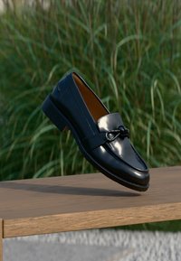 Μαύρο δερμάτινο μοκασίνι με διακοσμητικό κόμπο, που αιωρείται πάνω από ξύλινη επιφάνεια με ψηλό πράσινο χορτάρι στο φόντο.
