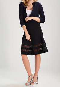 Granatowy kardigan koronkowy na białej bluzce, w połączeniu z czarną spódnicą z przezroczystymi koronkowymi paskami i srebrnymi sandałami na wysokim obcasie.