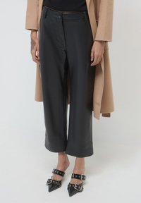 Pantaloni larghi in finta pelle nera con una finitura liscia, caratterizzati da un'alta vita e un cappotto beige a contrasto. Le calzature includono tacchi alti neri appuntiti con cinturini.