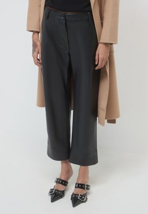 Schwarze, aus Kunstleder gefertigte weite Hosen mit glatter Oberfläche, hoch geschnitten, kombiniert mit einem kontrastierenden, kastanienbraunen Mantel. Die Fußbekleidung besteht aus spitzen schwarzen Heels mit Riemen.