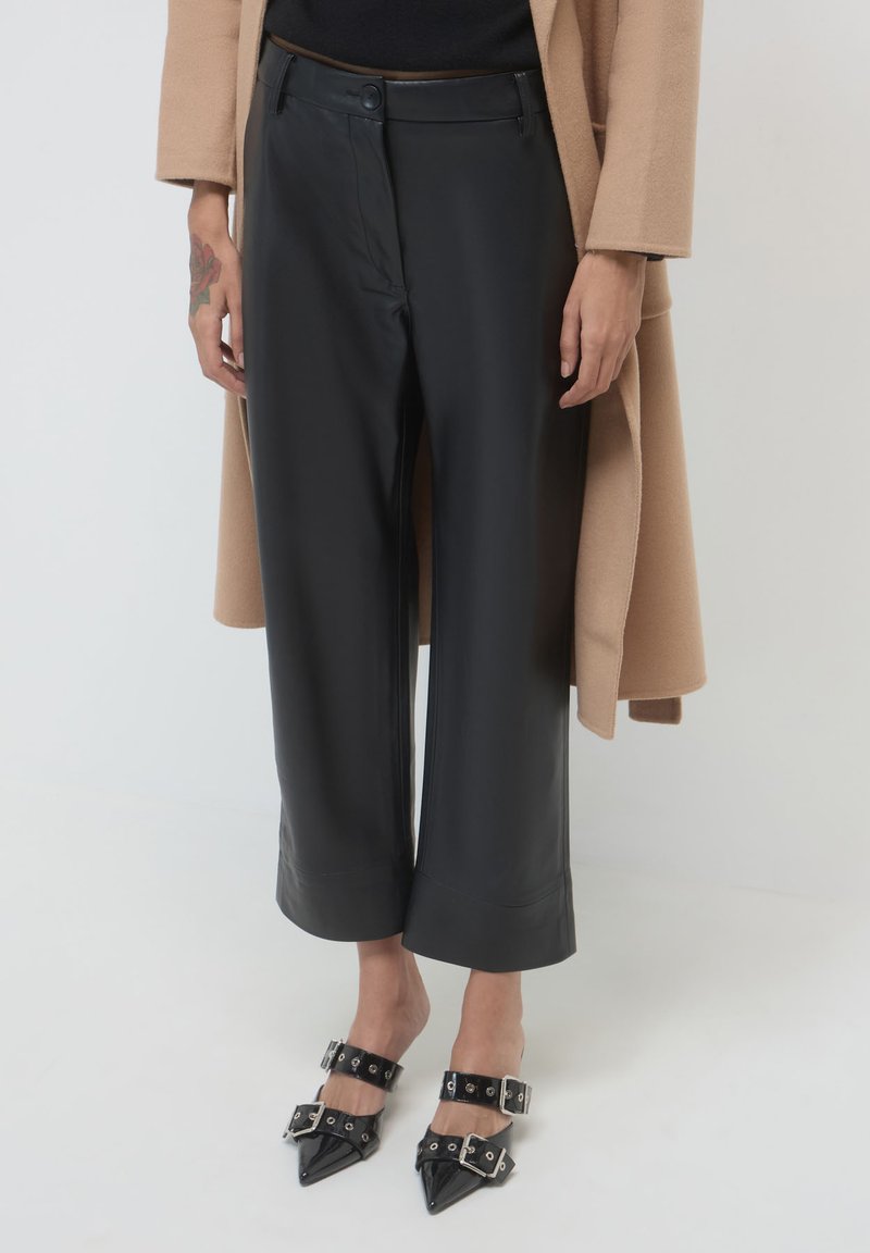 Pantaloni larghi in finta pelle nera con una finitura liscia, caratterizzati da un'alta vita e un cappotto beige a contrasto. Le calzature includono tacchi alti neri appuntiti con cinturini.