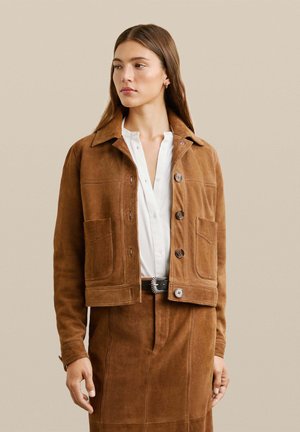 Bruine suède jas met een cropped ontwerp, grote voorzakken en knoopsluiting. Gecombineerd met een witte blouse en een bruine rok.