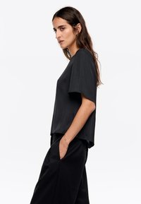Czarny top z krótkim rękawem, wykonany z gładkiego materiału o luźnym kroju, z okrągłym dekoltem i subtelnym rozcięciem po bokach u dołu.