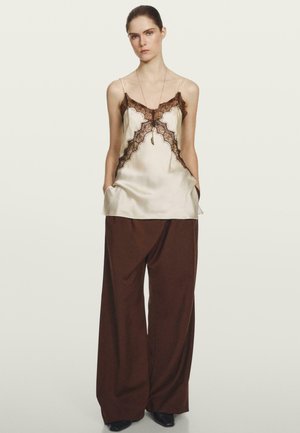 Satin-Camisole in hellem Beige mit schwarzem Spitzenbesatz, kombiniert mit weiten braunen Hosen. Das Outfit hat einen lockeren Sitz und tiefe Taschen.