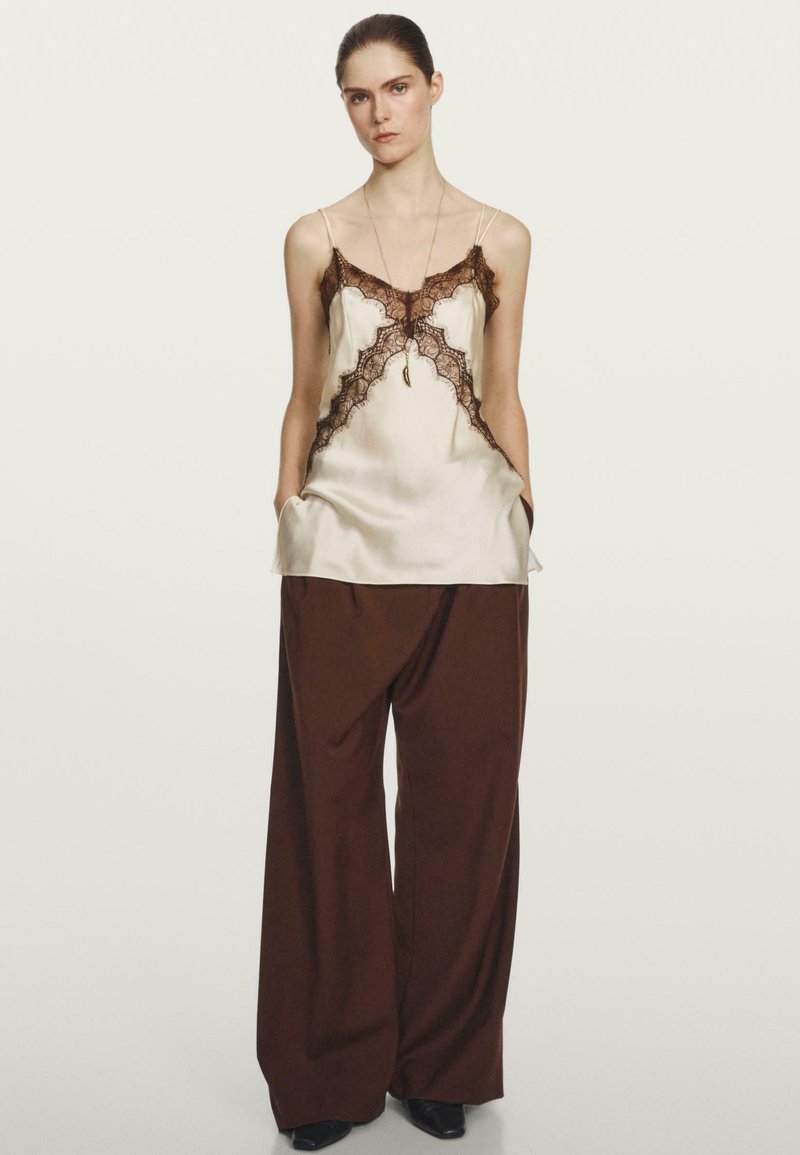 Satin-Camisole in hellem Beige mit schwarzem Spitzenbesatz, kombiniert mit weiten braunen Hosen. Das Outfit hat einen lockeren Sitz und tiefe Taschen.