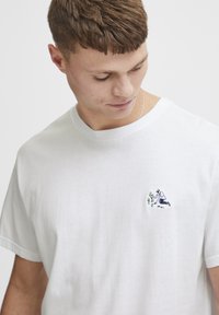 Hvid bomulds t-shirt med et lille broderet motiv af en person, der passer planter, på venstre bryst. Rund halsudskæring, korte ærmer.