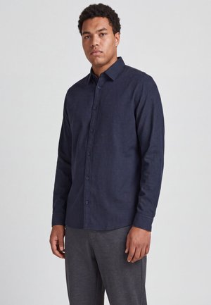 Homme portant une chemise boutonnée bleu foncé et un pantalon gris, debout devant un fond gris clair uni.